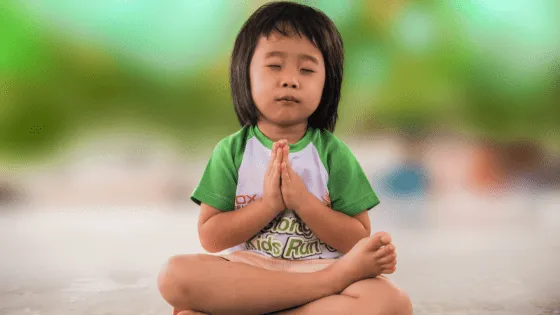 Criança sentada de pernas cruzadas, de olhos fechados e mãos unidas em meditação