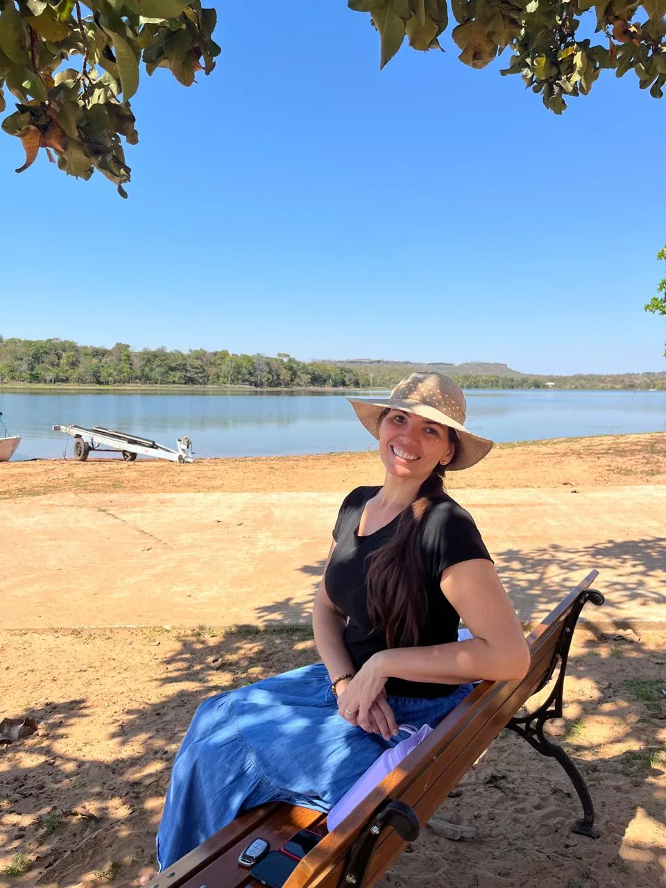 Elicéia Trindade de chapéu, sentada em um banco à beira de um lago em dia de sol